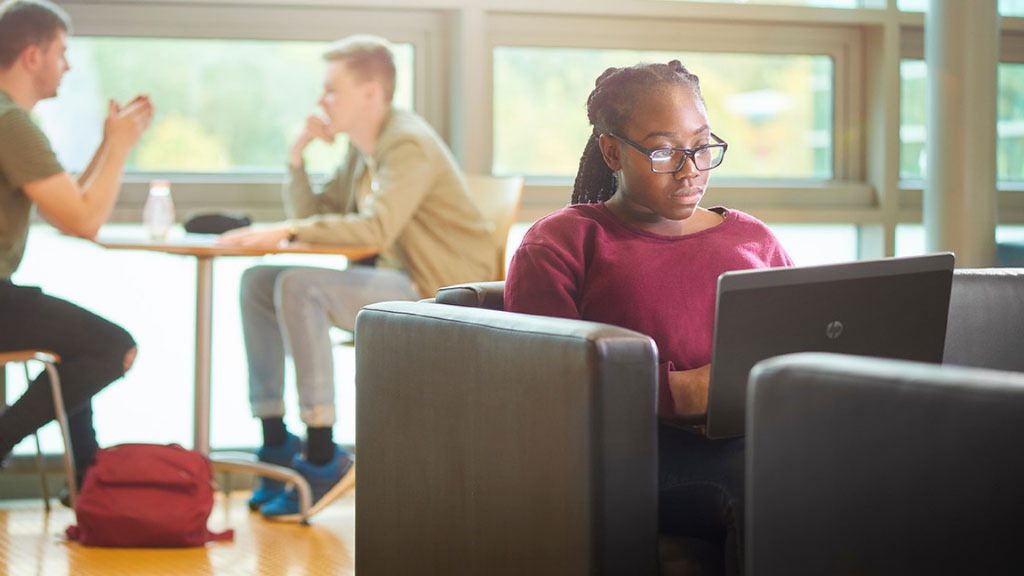 An image of a student sat on a couch holding a laptop, in the background of the image there are two people sat at a small table.