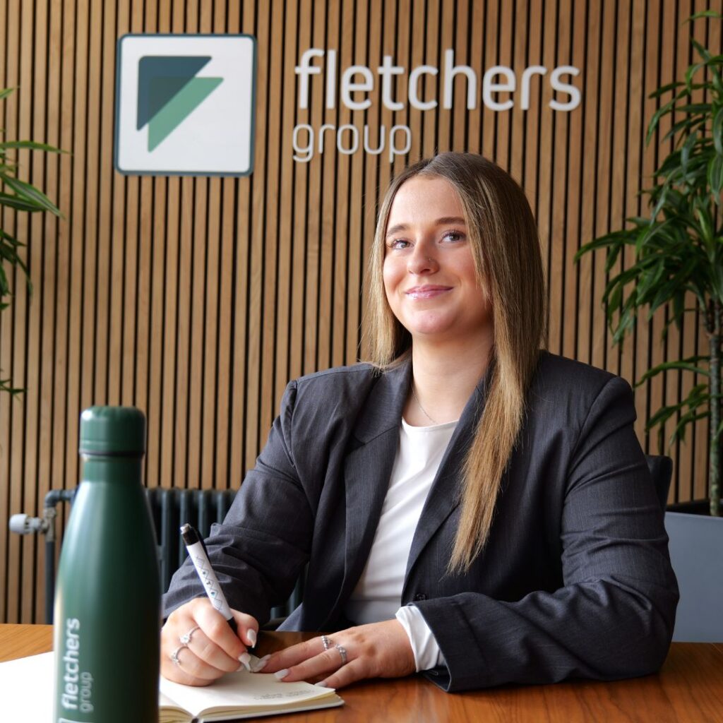 Photo of Emily Forster sat behind a desk at Fletchers Solicitors with the company logo on the wall behind her
