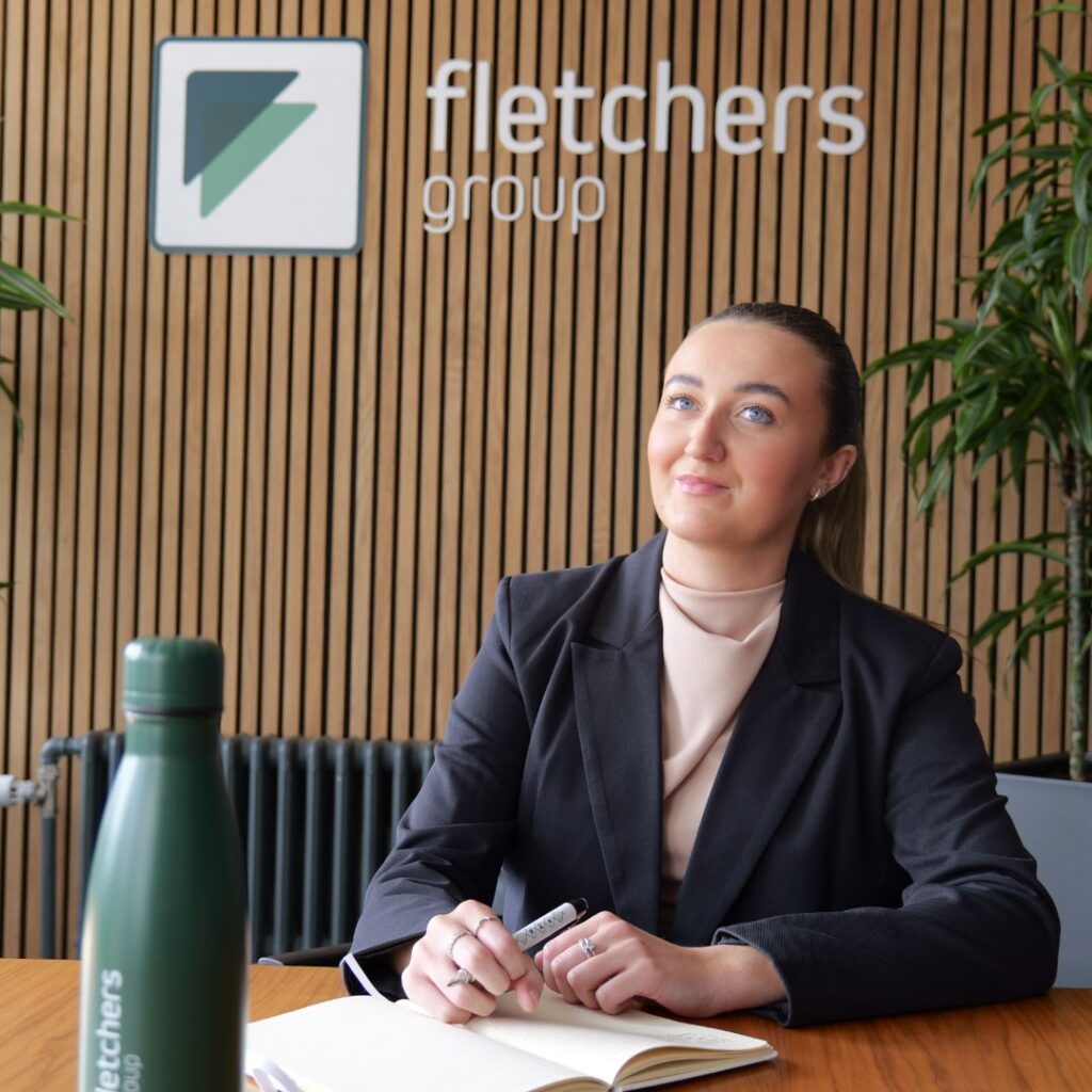 Photo of Freya Bulcock sat behind a desk at Fletchers Solicitors with the company logo on the wall behind her