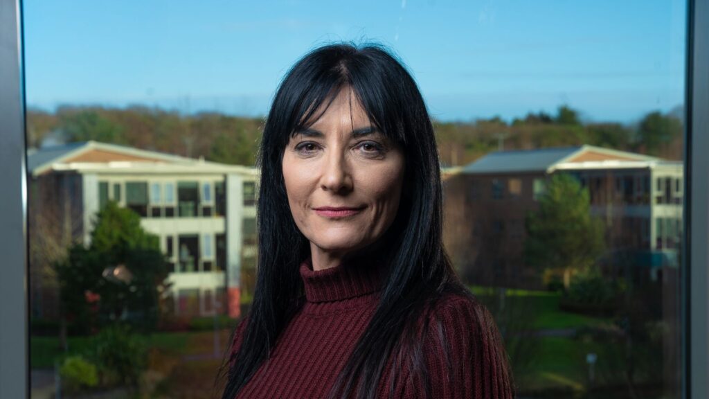 Landscape photo of Abigail Moriarty standing in front of a window looking straight at camera smiling, wearing a maroon turtleneck jumper.