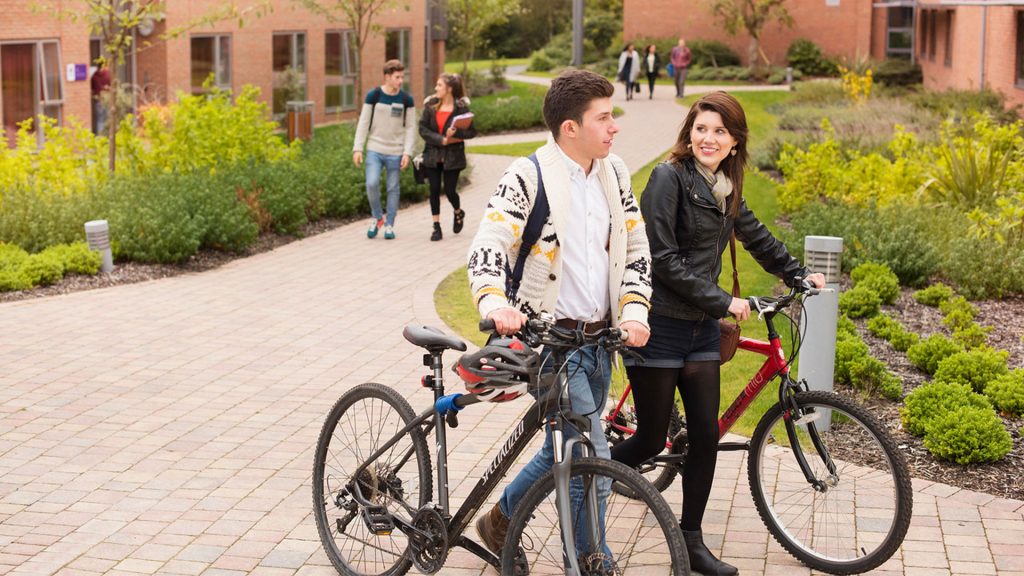 Two students walking together past accommodation with their bikes. Other students are walking past in the background.