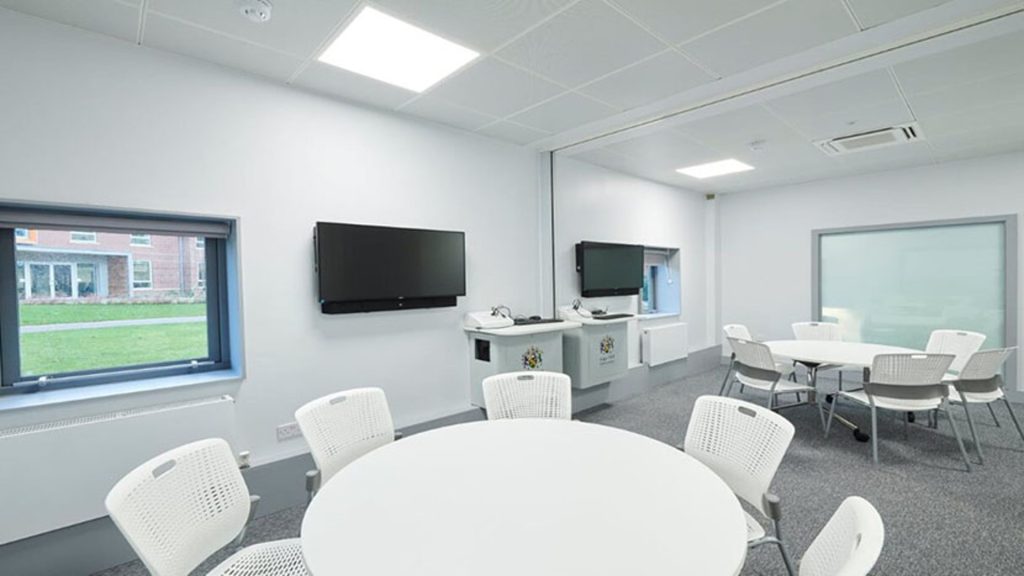 White chairs are surrounding a white table in the foreground. There are TVs on the wall in the background. The room is white.