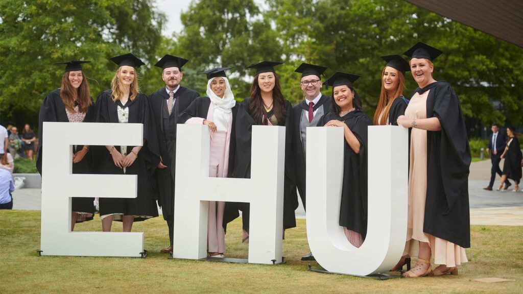 A group of graduates stand behind an 'EHU' sign after a graduation ceremony