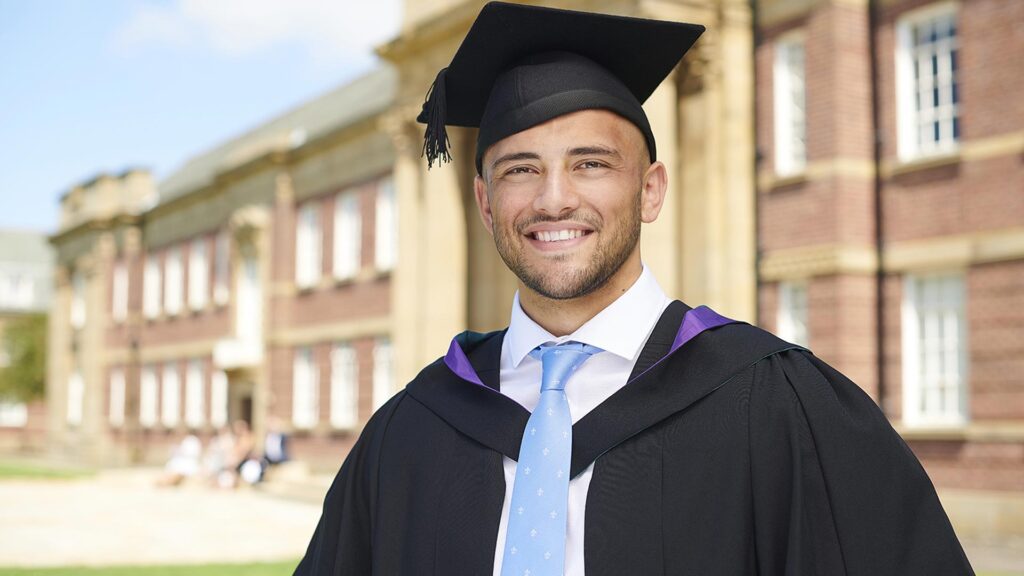 A graduate stands outside the front of the Main Building wearing their cap and gown.