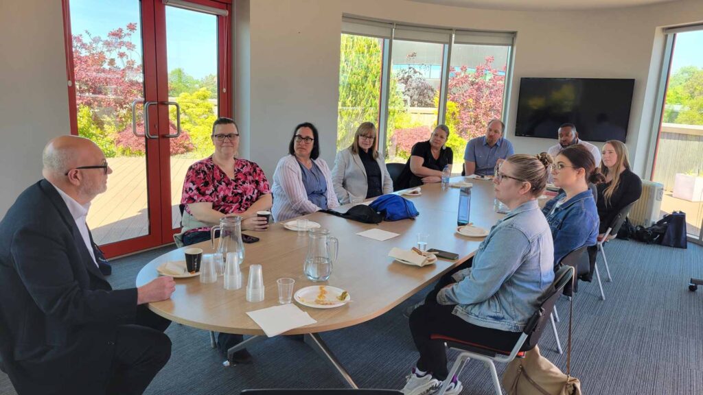 12 visitors from the University of Toledo sitting in a board room listening to staff  male leader from Edge Hill University 
