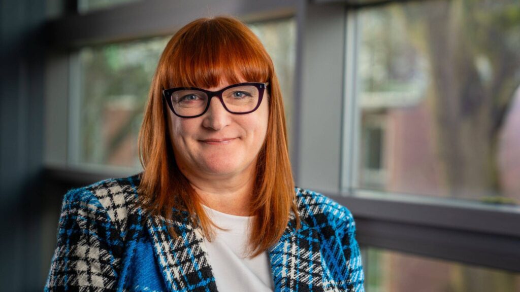 Dr Jo Wright stands in front of windows and smiles at the camera